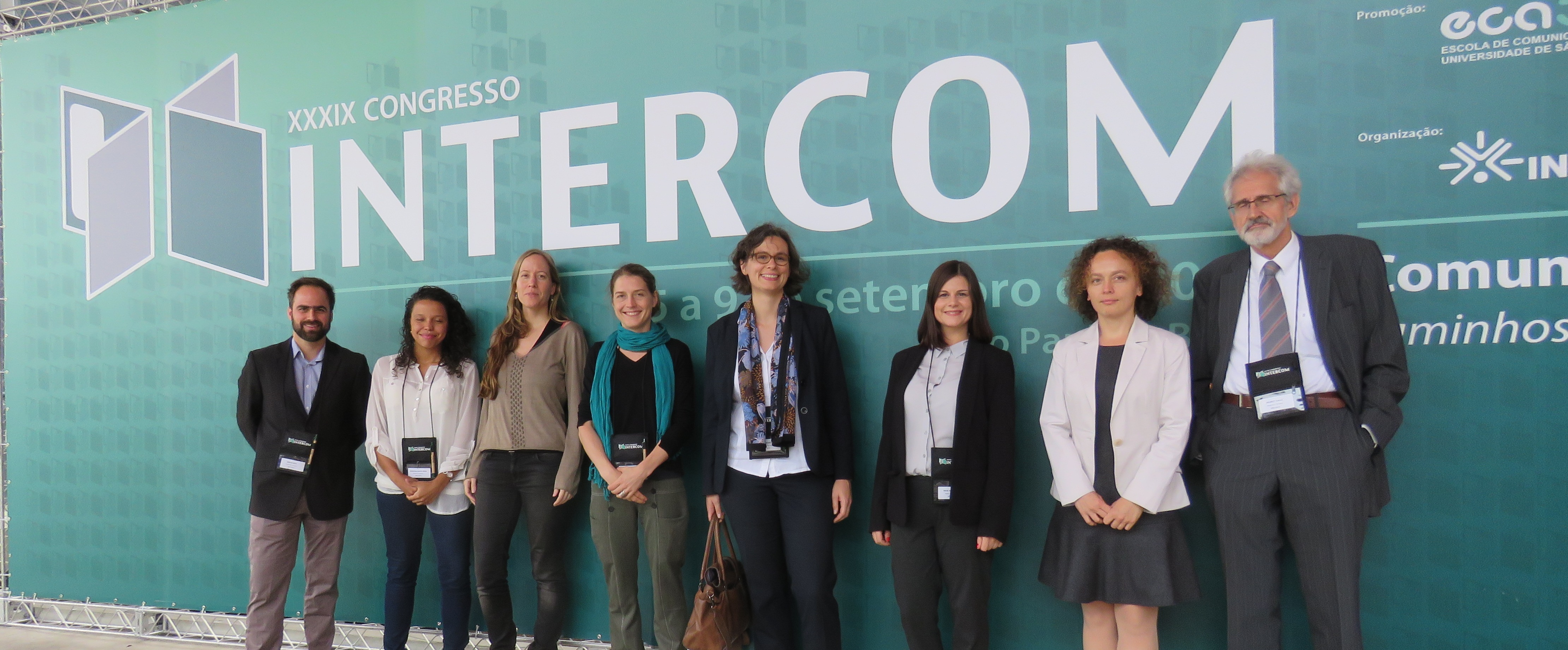 Oito representantes (homens e mulheres) de instituições alemãs se posicionam em frente a um banner com o logotipo da INTERCOM