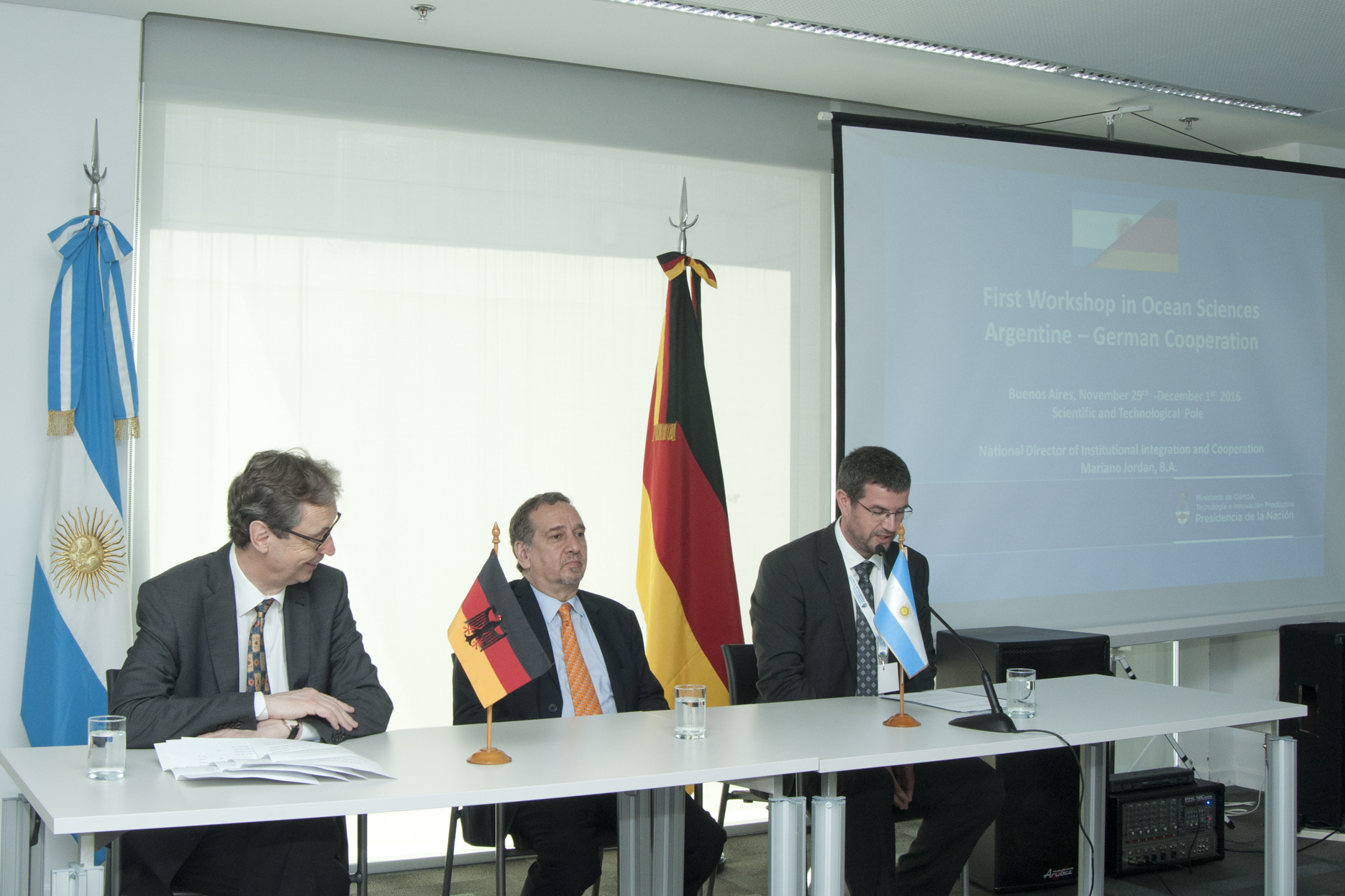 A imagem mostra as três autoridades sentadas à mesa com as bandeiras da Argentina e da Alemanha ao fundo. Do lado direito da imagem está um telão com projeção do workshop