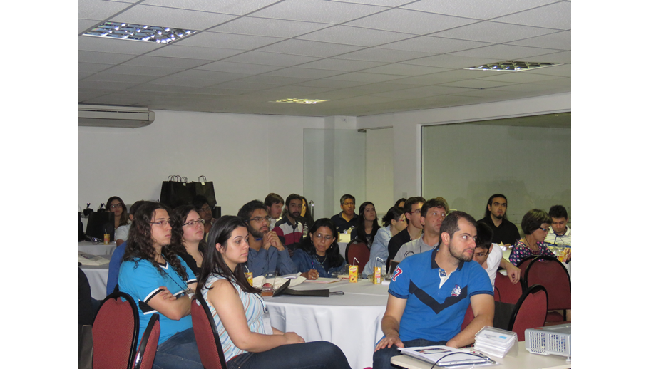 A imagem mostra um grande número de participantes sentados em diferentes mesas e atentos a uma palestra