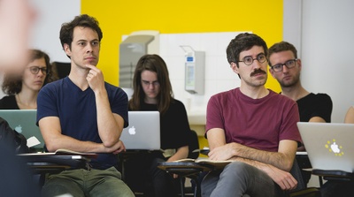 A imagem mostra pessoas sentadas com computadores prestando atenção em algo