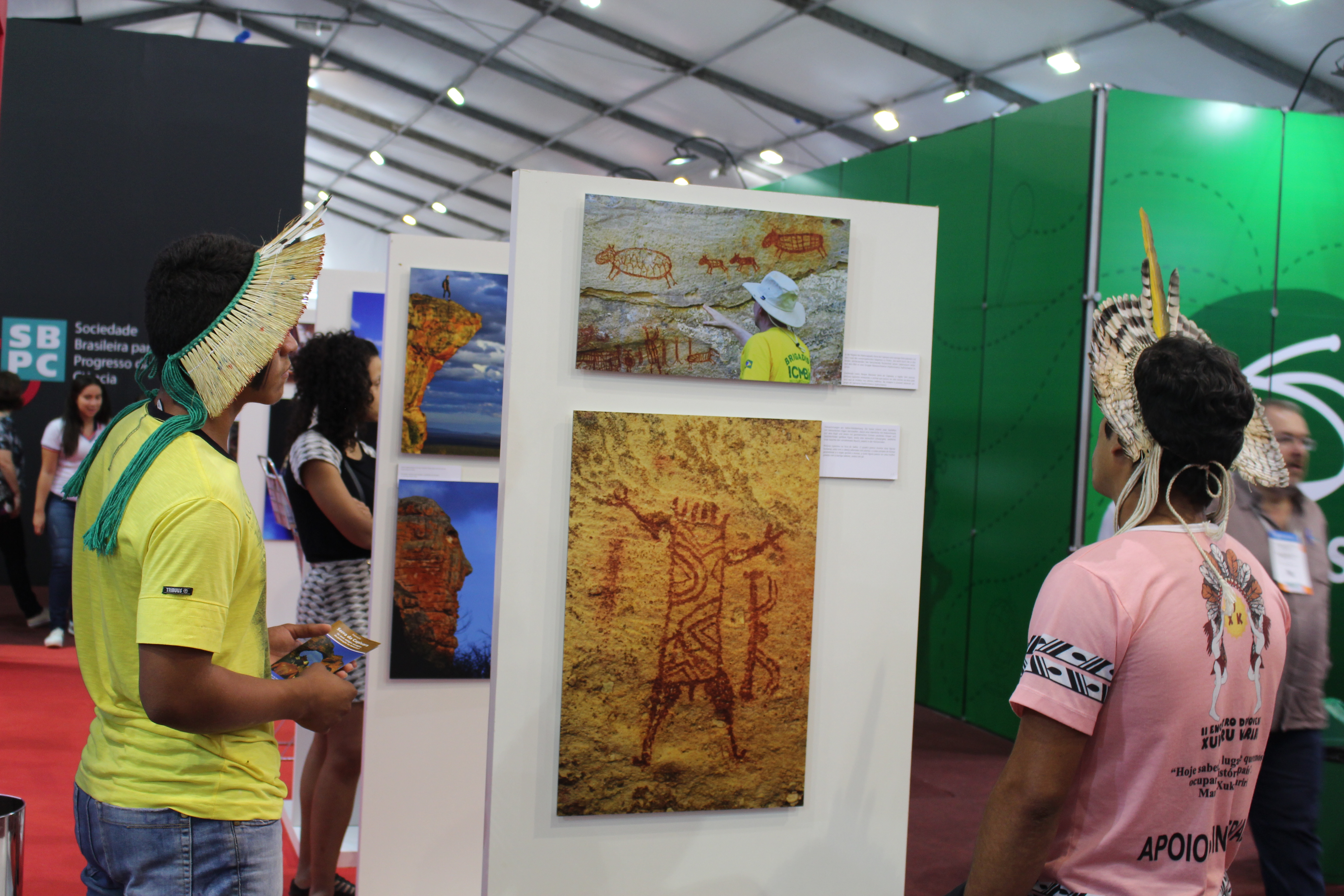 Dois visitantes usando cocares indígenas apreciam fotos de pinturas rupestres da exposição