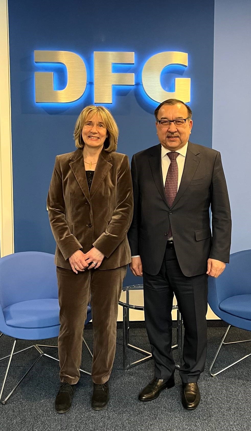 H. E. Dr. Nurlan Onzhanov and Dr. Heide Ahrens stand next to each other in front of an illuminated DFG sign, two chairs and a small table and pose for the photo.