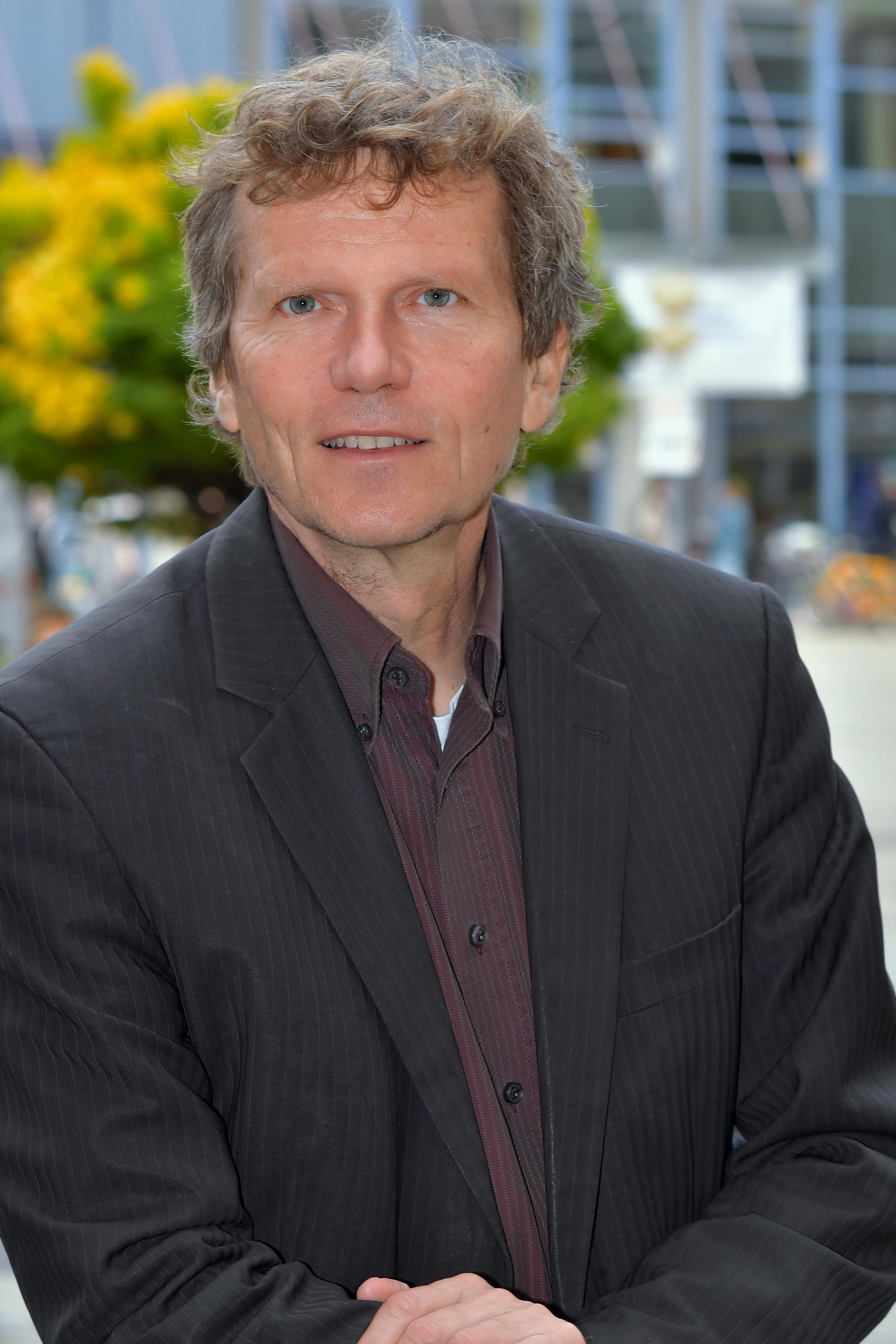 Prof. Dr. Hartmut Rosa steht im Anzug vor einem blauen Gebäude und grünen und gelben Pflanzen.