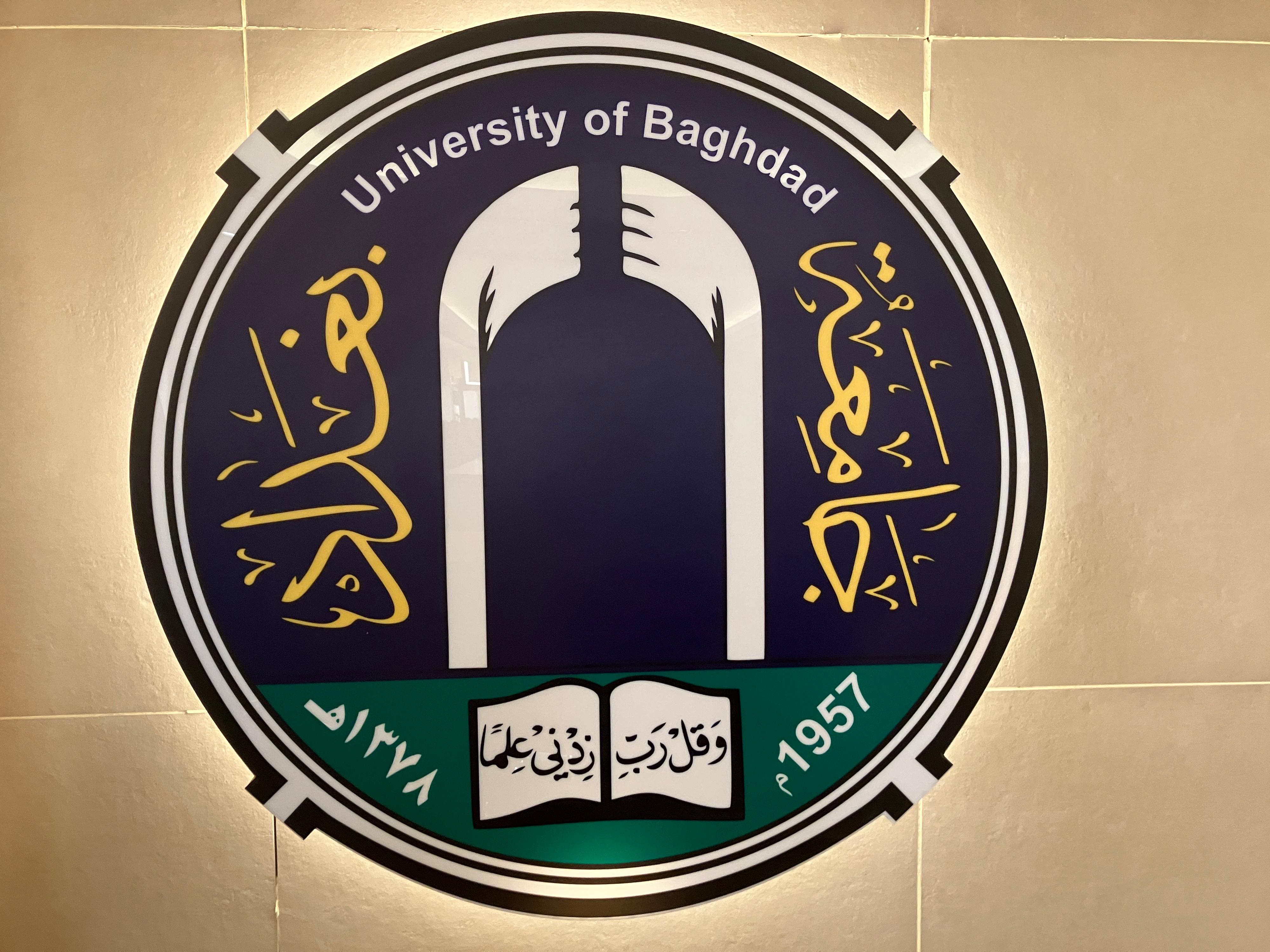Das runde blau-grüne Logo besteht aus goldener arabischer Schrift an den Seiten und unten, außerdem steht in Englisch "University of Bagdad" oben und "1957" rechts unten.