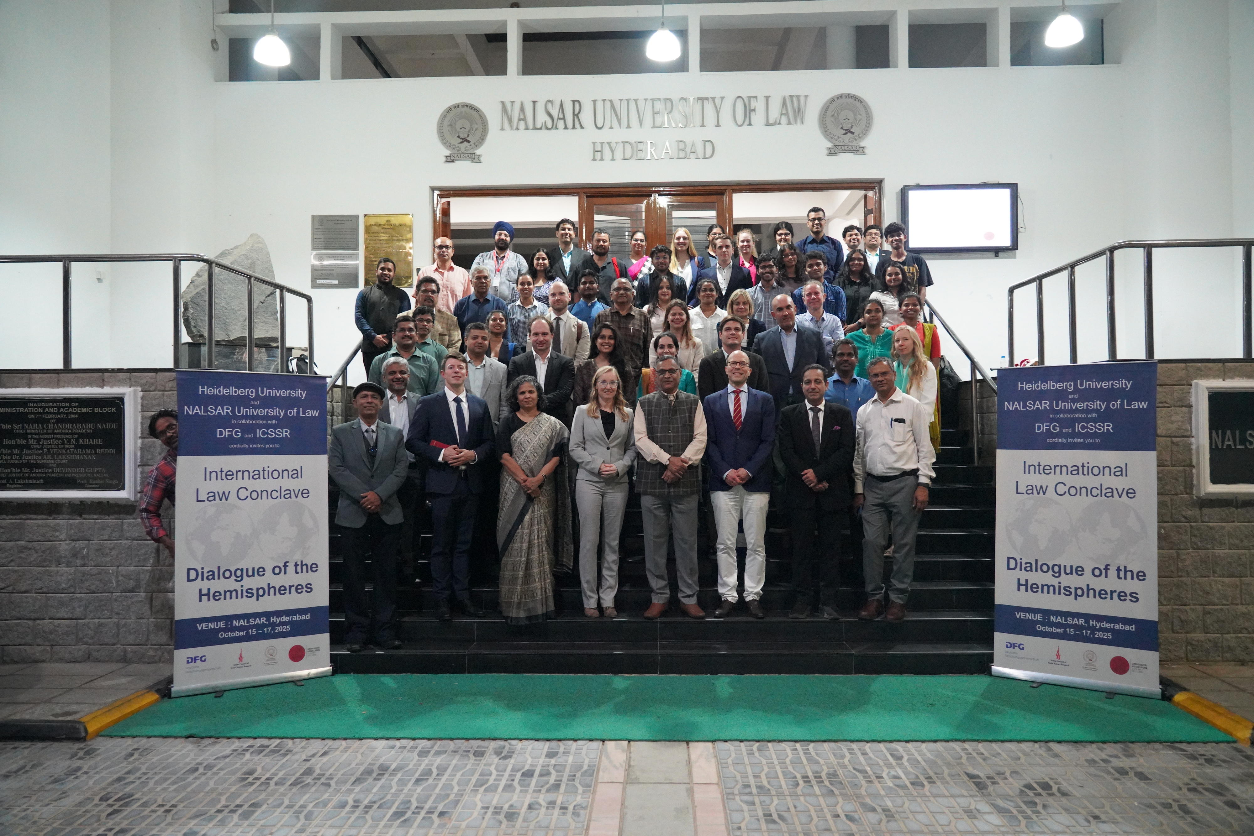 Group picture in front of the NALSAR University of Law, on the right and left side posters of the event.