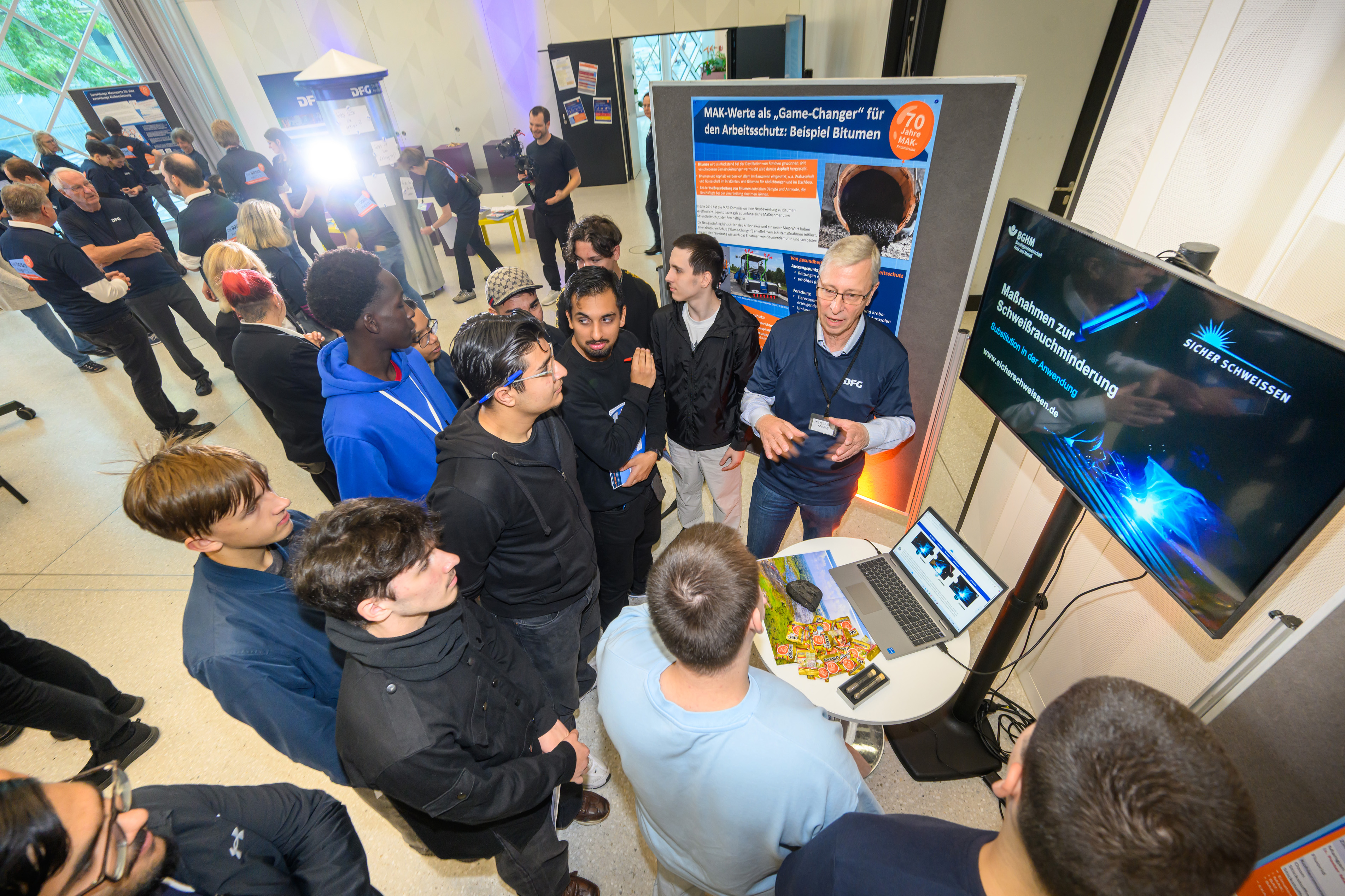 An einem Stand der Ausstellung spricht ein Experte zu einer Gruppe junger Menschen vor einem Computer und einem Bildschirm 