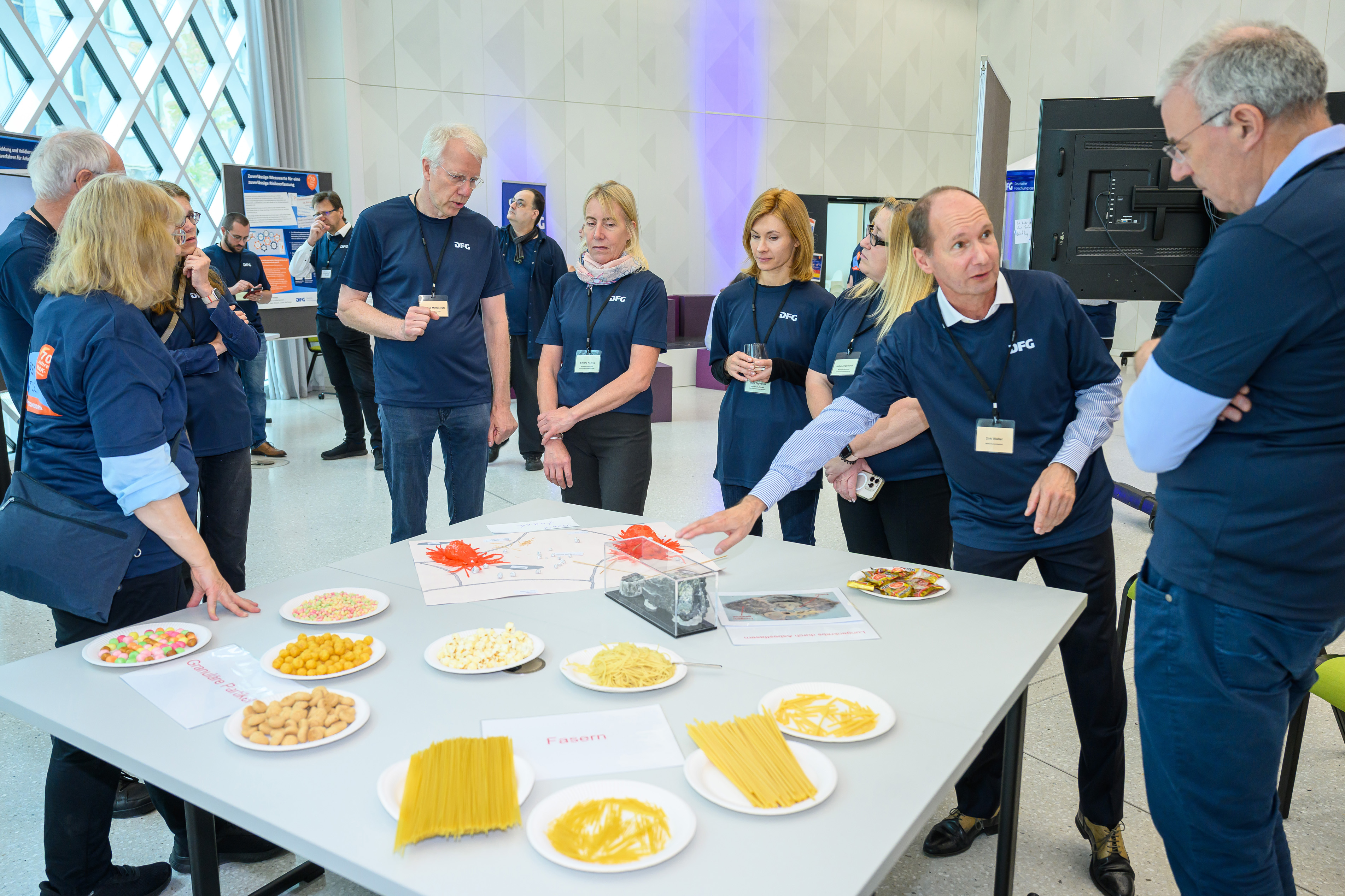 Menschen stehen um einen Tisch, auf dem Lebensmittel stehen, die die Form von Partikeln illustrieren, und wie sich dies auf den menschlichen Körper auswirkt