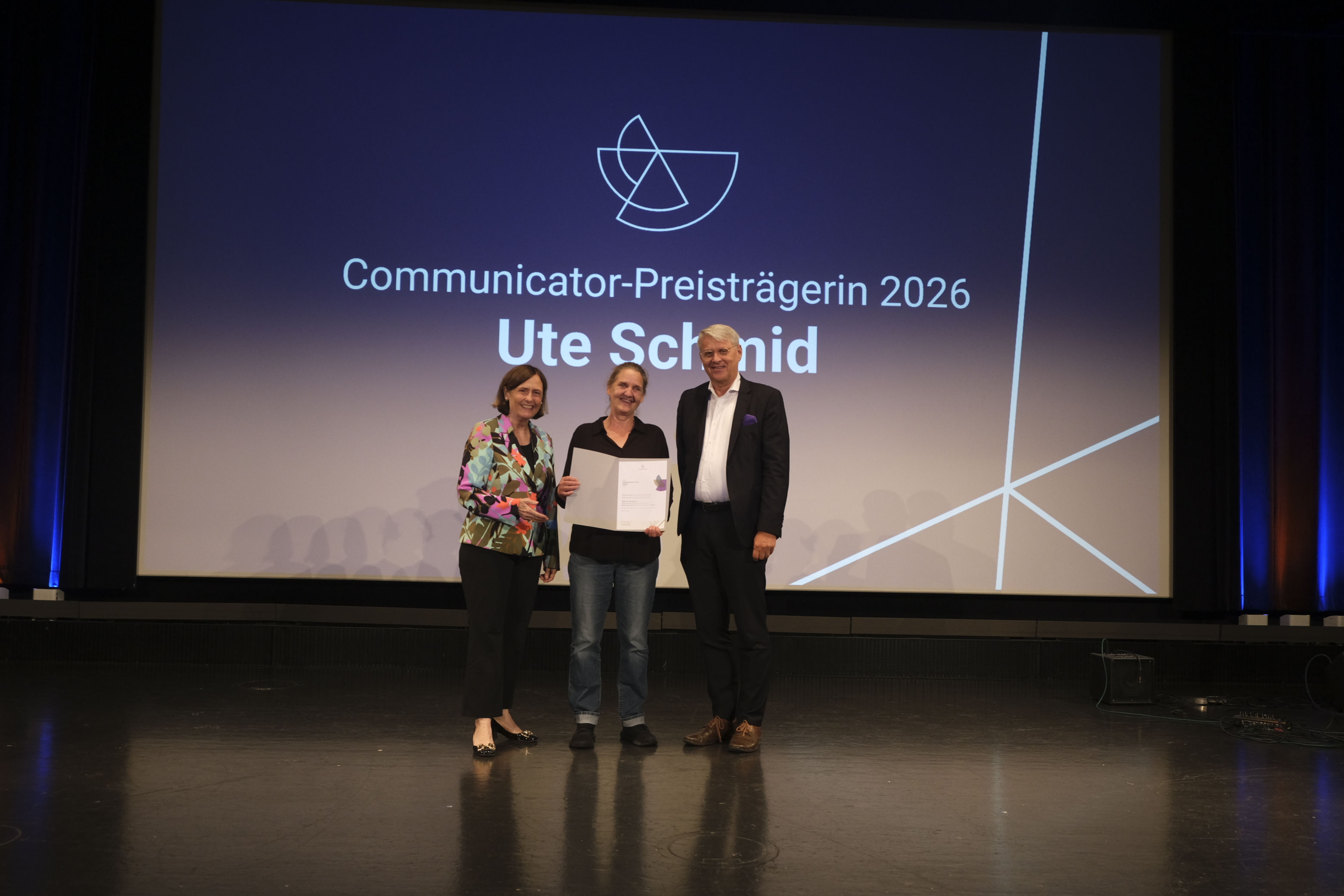 Die Urkunde des Communicator-Preis 2026 für herausragende Wissenschaftskommunikation übergeben die Präsidentin der DFG, Prof. Dr. Katja Becker und der Generalsekretär des Stifterverbandes, Volker Meyer-Guckel, gemeinsam an die Preisträgerin Prof. Dr. Ute Schmid (Professorin für Kognitive Systeme, Universität Bamberg)