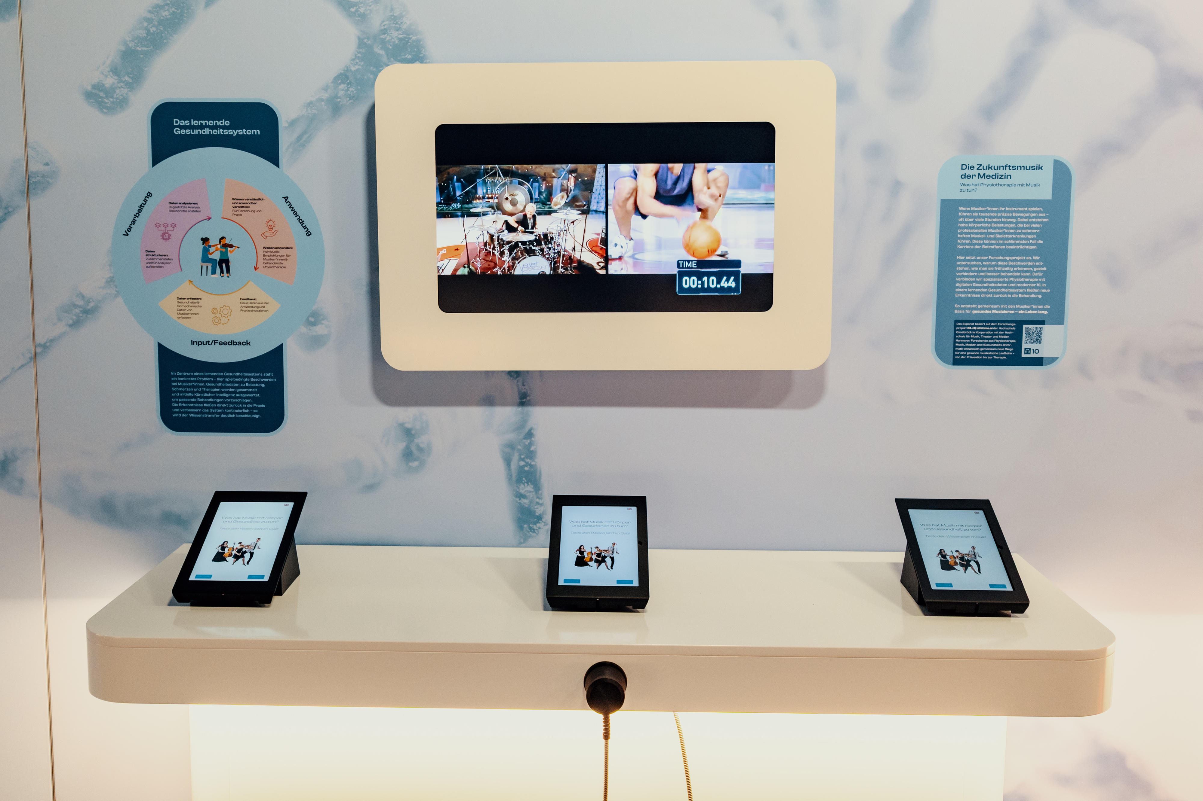 Bild eines Ausstellungstisches mit drei Tablets, darüber hängt mittig an der Wand ein Bildschirm, links und rechts davon jeweils ein Infotext