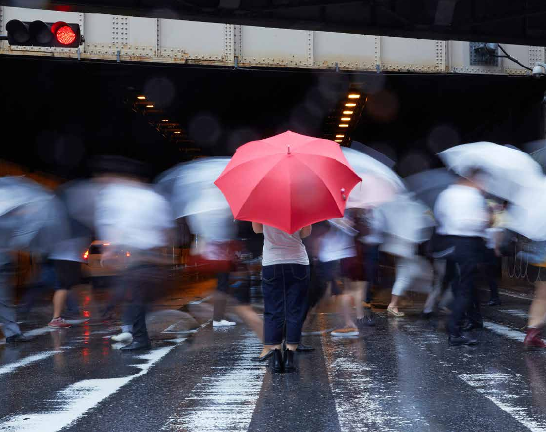 Bild eines Fußgängerüberwegs mit vielen Personen, die in Bewegung sind und Regenschirme halten. Mittig steht eine Frau mit einem roten Regenschirm.