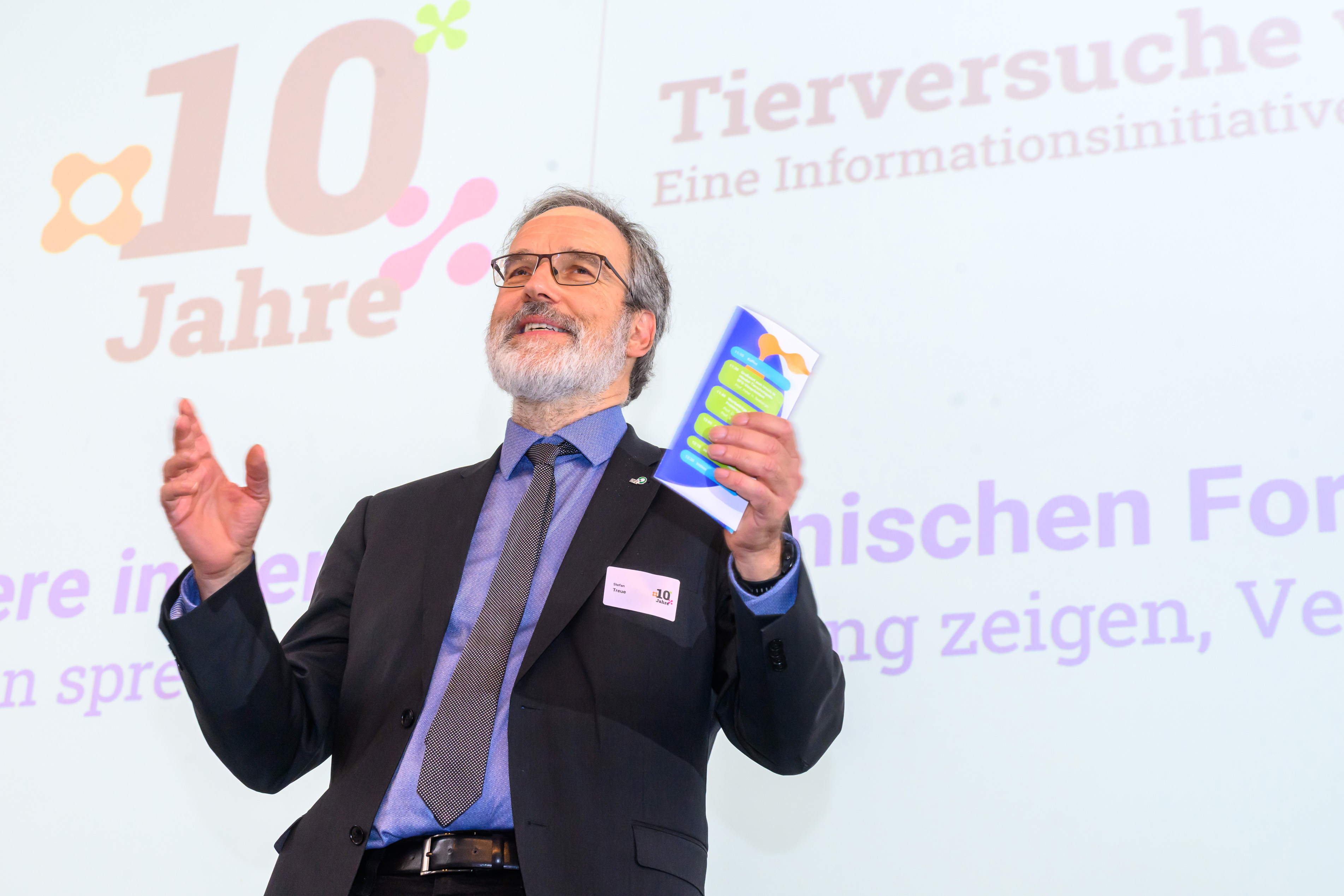 Stefan Treue steht auf der Bühne und spricht mit dem Veranstaltungsprogramm in der Hand zu den Teilnehmer*innen
