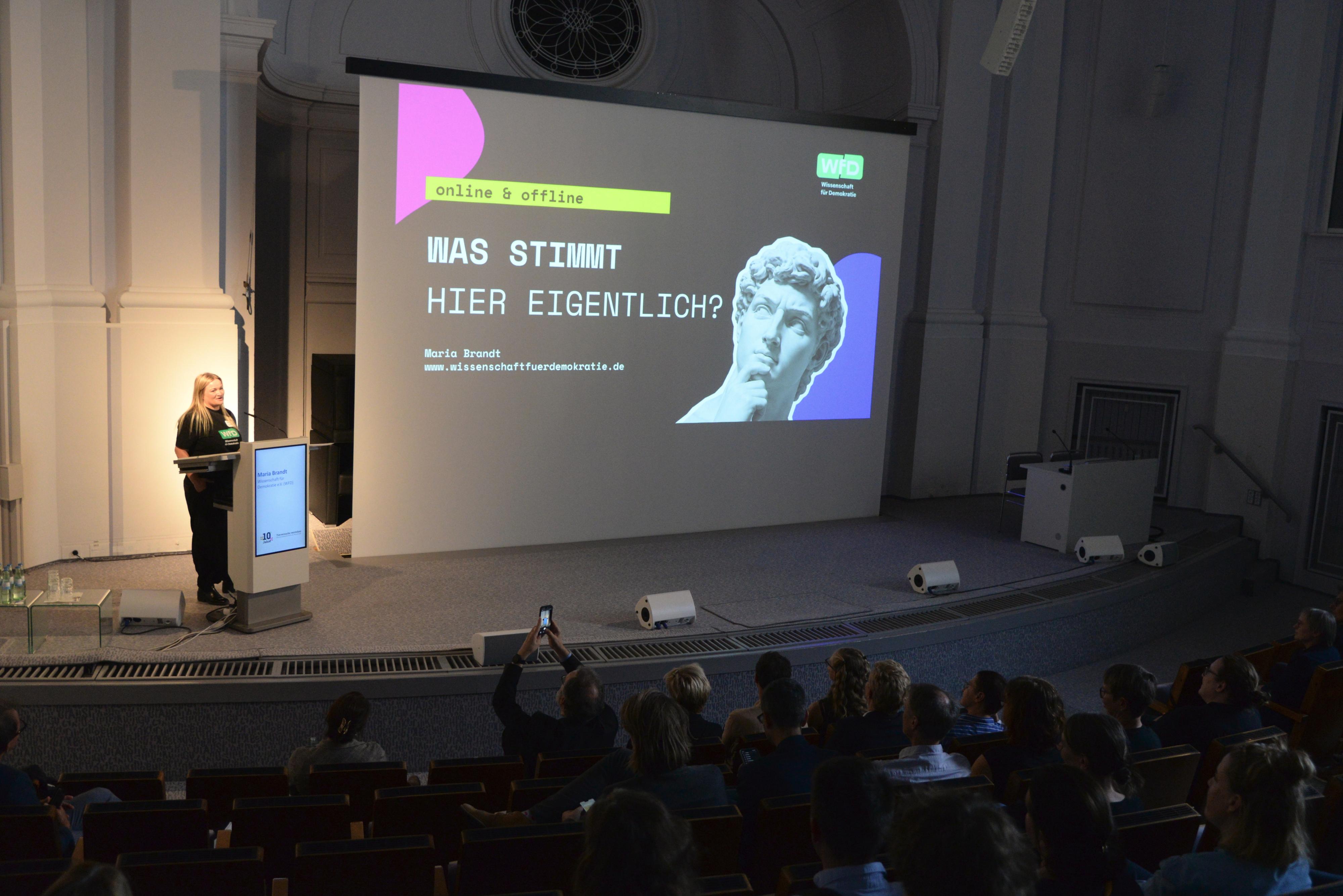 Maria Brandt steht vor einer Präsentation, auf der steht "Was stimmt eigentlich?", auf der Bühne